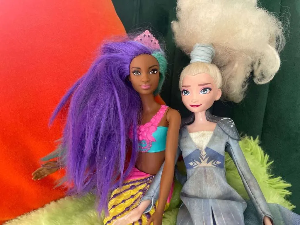 Black mermaid barbie with purple hair and Elsa barbie in clue dress sitting on green and orange cushions.